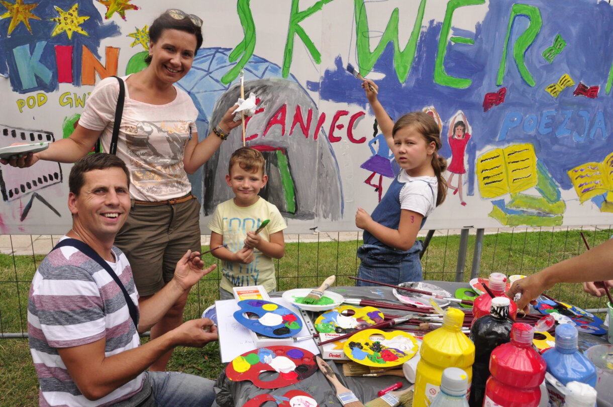 Dwoje dorosłych i dwoje dzieci przy stoliku z farbami, w tle pomalowana ścianka