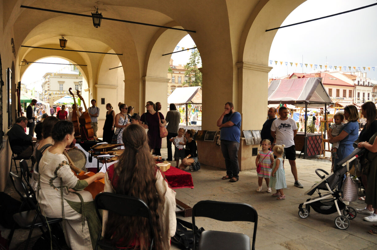 Trzy grające na instrumentach kobiety w podcieniach, w tle gapiowie