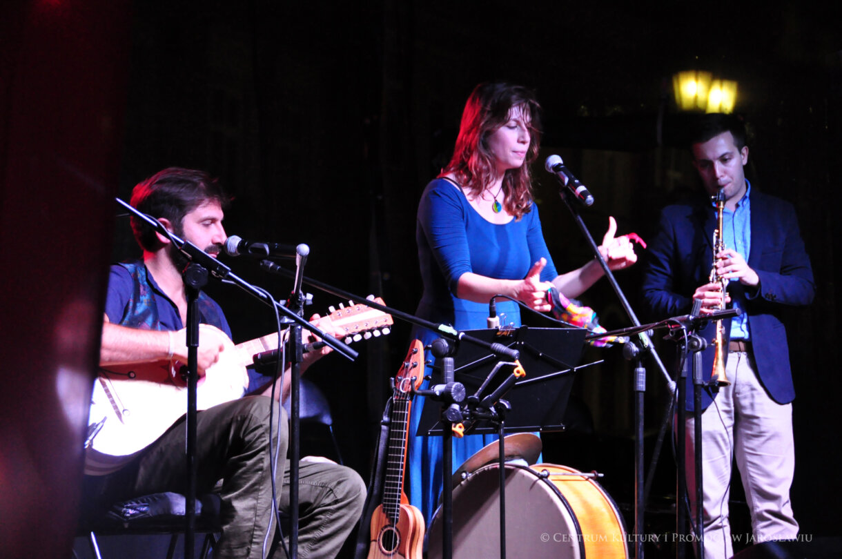 Mężczyzna grający na gitarze, kobieta śpiewająca, w tle mężczyzna grający na klarnecie