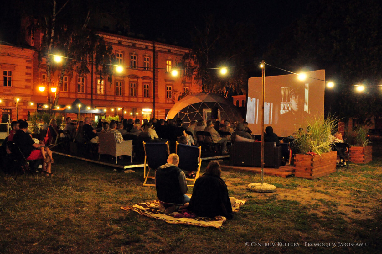 Z przody dwie osoby siedzące na kocu, w tle ludzie oglądający seans kina plenerowego