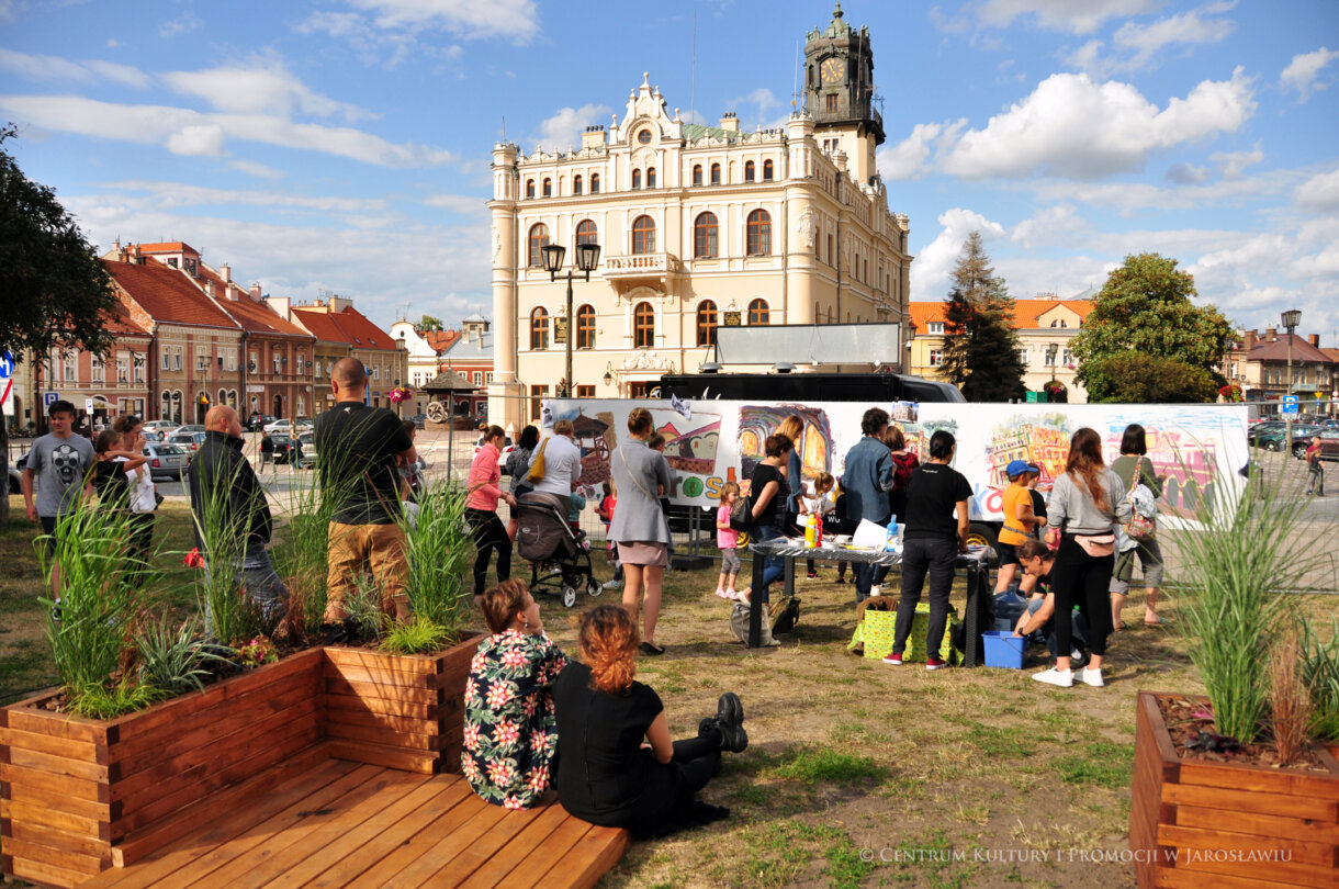 Z przodu - dwie osoby siedzące na podeście, z tyłu grupa ludzi malująca na ściance