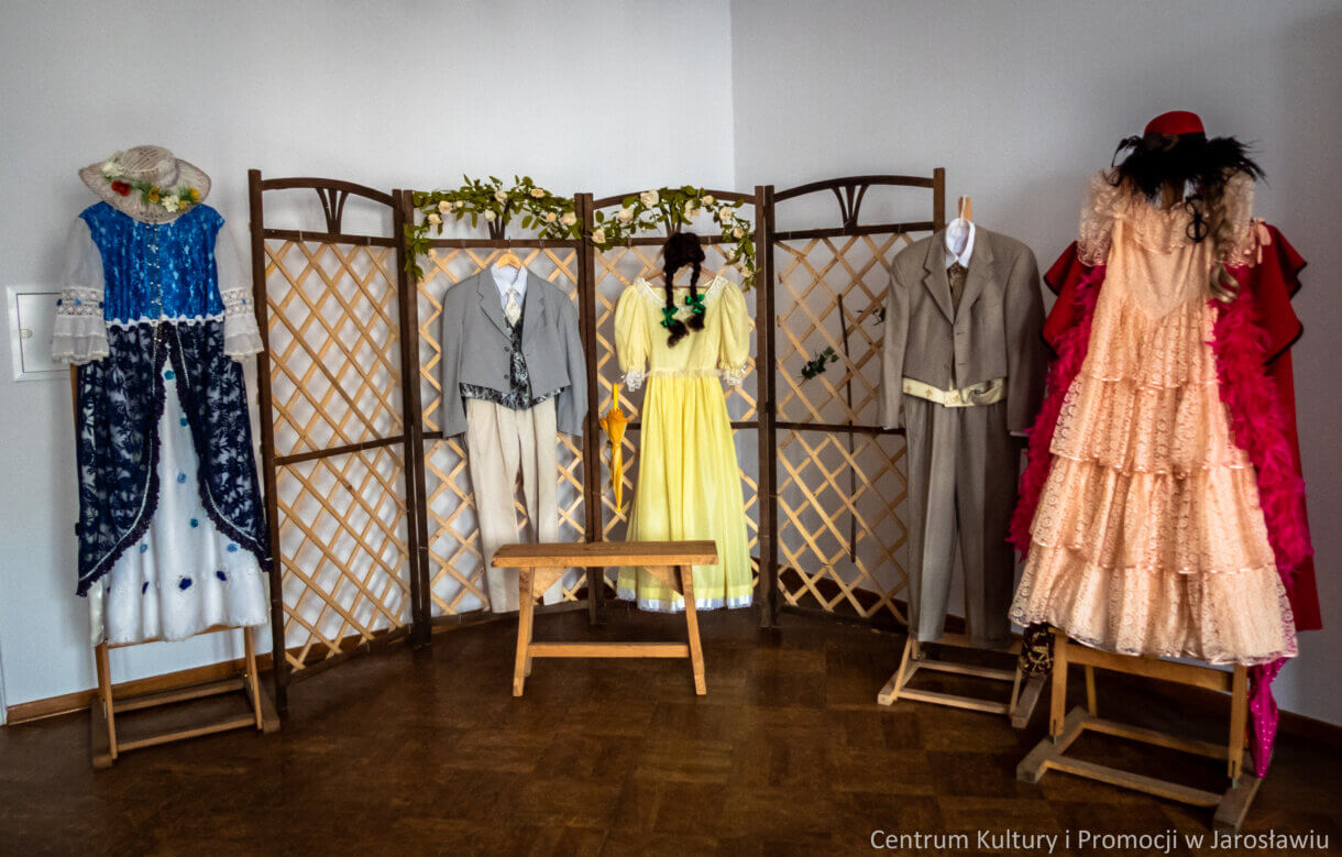 scenografia do sztuki, niebiesko-biała suknia, letni jasny męski garnitur, żółta suknia z czarną peruką, szary garnitur, różowa sukienka z czerwonym płaszczem. Przed pergolą ustawiono ławkę