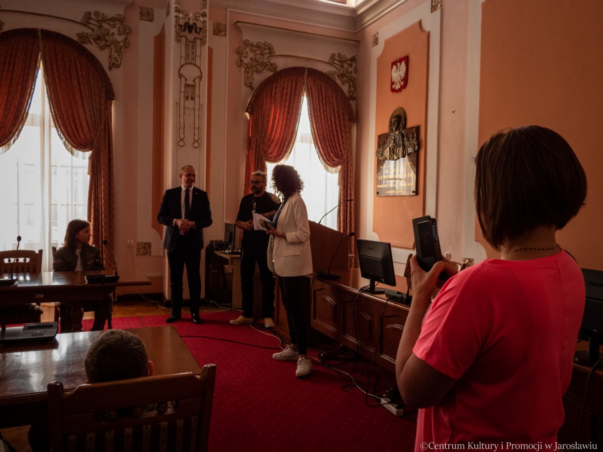 zwiedzanie Sali Narad, w tle zastępca burmistrza miasta, radny rady miasta oraz przewodnik po ratuszu